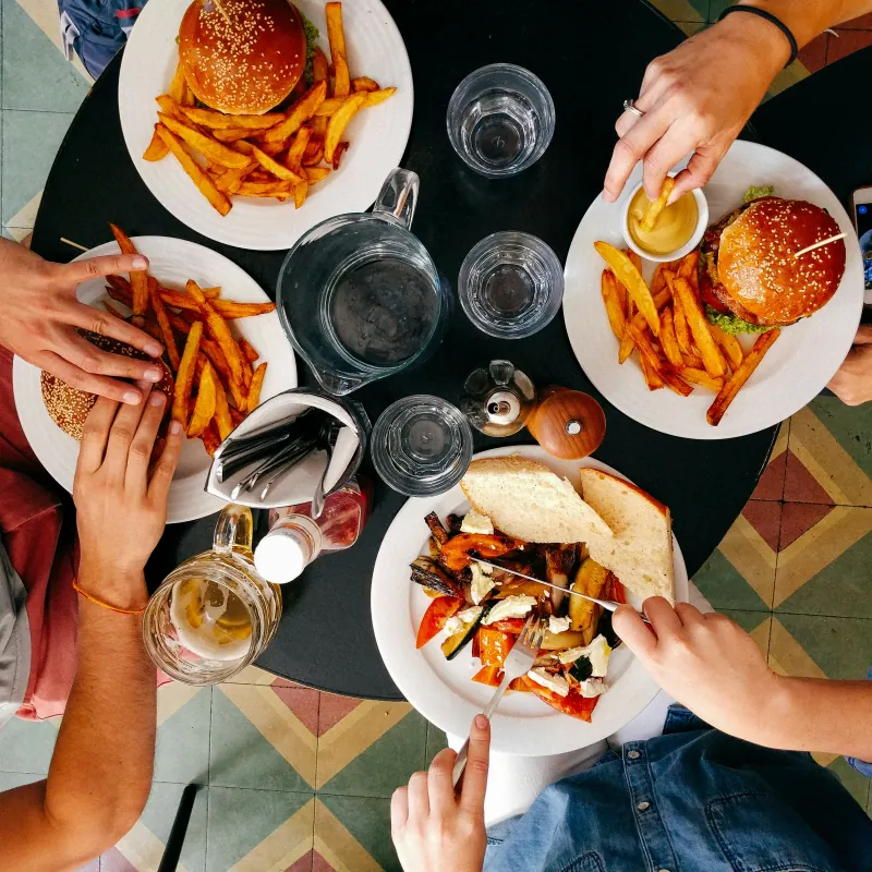Personas comiendo hamburguesas con patatas fritas y verduras asadas, vistas desde arriba, en una mesa redonda